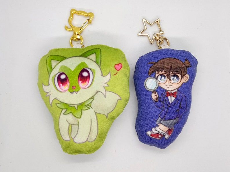 May include: Two plush keychains. One is green and features a cartoon white fox with red eyes. The other is blue and features a cartoon boy with brown hair wearing a blue jacket and holding a magnifying glass.