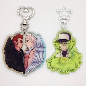 May include: Two acrylic keychains with printed images. One keychain features a heart-shaped design with two men, one with black wings and the other with white wings. The other keychain features a green-haired character with a white hat and a white shirt.