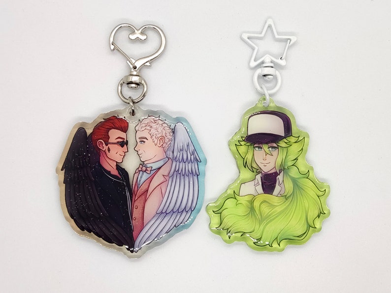 May include: Two acrylic keychains with printed images. One keychain features a heart-shaped design with two men, one with black wings and the other with white wings. The other keychain features a green-haired character with a white hat and a white shirt.