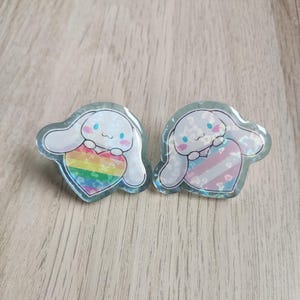 May include: Two iridescent pins featuring a cartoon character holding a heart. One heart is rainbow coloured and the other is a transgender pride flag.