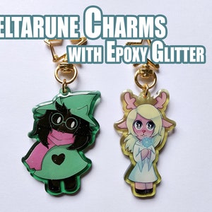 Könnte beinhalten: Zwei Deltarune-Anhänger mit Epoxid-Glitter. Der Anhänger links ist grün und zeigt eine Figur mit einem rosa Schal und einer Brille. Der Anhänger rechts ist weiß und zeigt eine Figur mit blonden Haaren und Geweih. Beide Anhänger haben goldene Sprungringe.