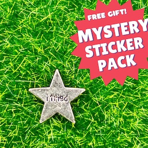 Può includere: Un ciondolo a forma di stella argentato con le parole "i tried" su di esso. Il ciondolo è su uno sfondo verde con un'esplosione di stelle rossa sullo sfondo che dice "REGALO GRATUITO! PACCHETTO DI STICKER MISTERO".