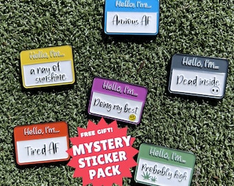 Op de afbeelding: Zes emaillepins met verschillende spreuken. De pins hebben allemaal de vorm van een naamschild en een zwarte rand. De spreuken op de pins zijn "Hello, I'm... a ray of sunshine", "Hello, I'm... Anxious AF", "Hello, I'm... Tired AF", "Hello, I'm... Doing my best", "Hello, I'm... Dead inside", en "Hello, I'm... Probably high". De pins bevinden zich allemaal op een groene ondergrond. In het midden van de afbeelding bevindt zich een rode ster met de tekst "FREE GIFT! MYSTERY STICKER PACK".