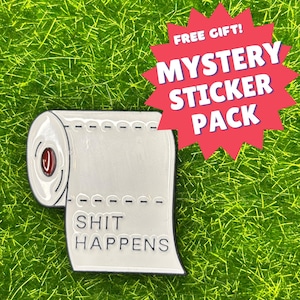 Könnte beinhalten: Weißer Emaille-Pin in Form einer Toilettenpapierrolle mit dem schwarzen Schriftzug „SHIT HAPPENS“. Der Pin hat eine rote Mitte und ist vor einem grünen Hintergrund platziert. Eine rote Sternburst-Grafik lautet „FREE GIFT! MYSTERY STICKER PACK“.