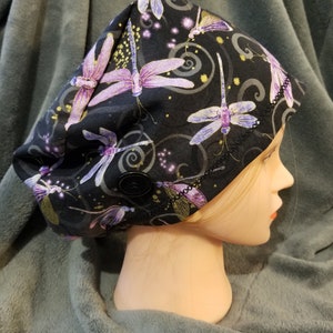 May include: Black scrub hat with a purple and gold dragonfly print. The hat has a button on the side.