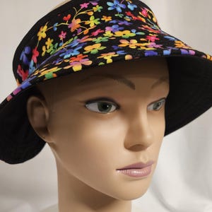 May include: A black sun visor featuring a vibrant, multi-colored floral pattern with small flowers in shades of red, orange, yellow, green, blue, and purple. The visor has a wide, curved brim and a solid black underside, providing full coverage.