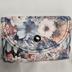 May include: A small, floral-patterned wallet with a snap closure. The wallet features a design of pink, white, and blue flowers on a textured background. The flap is secured with a silver-colored snap.