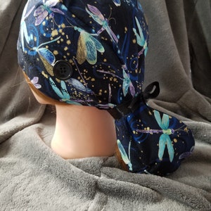 May include: A black scrub hat with a blue and gold dragonfly print. The hat has a button on the side and a long tie.