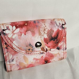 May include: A small, floral-patterned wallet with a snap closure. The wallet features a watercolor design with pink, red, and white flowers. The edges are stitched with red thread, and the snap is silver.