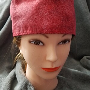 May include: A red scrub cap with a speckled pattern. The cap is made of fabric and is worn on a mannequin head. The mannequin has brown eyes and orange lipstick. The background is a gray blanket.