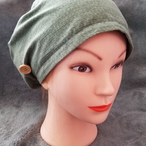 May include: A green slouchy beanie with a single button on the side. The beanie is made of a soft, stretchy fabric.