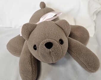 May include: A plush brown teddy bear with black button eyes and a black nose. The bear has a light pink ribbon tied in a bow around its neck. The bear is lying on a white surface.