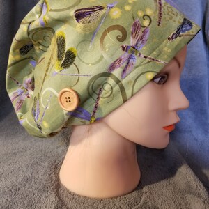May include: Green scrub hat with a dragonfly print and a brown button. The hat is on a mannequin head.