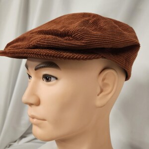 May include: A brown corduroy flat cap with a short brim. The cap is a solid color and has a ribbed texture. The cap is displayed on a mannequin head.