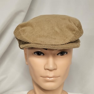 May include: A tan corduroy flat cap. The hat has a rounded crown and a short, stiff brim. The corduroy fabric has vertical ridges. The cap is a neutral color, suitable for various outfits. The hat is a classic style.
