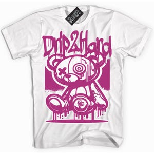 Könnte beinhalten: Weißes T-Shirt mit einem pinkfarbenen Aufdruck eines tropfenden Teddybären mit einem Auge und einem X über dem anderen. Der Text "Drip2Hard" befindet sich über dem Bären.
