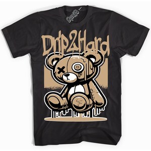 Könnte beinhalten: Schwarzes T-Shirt mit einem Cartoon-Teddybär-Design und dem Text "Drip 2 Hard". Der Bär ist braun, weiß und schwarz mit einem Knopfauge und einem X für das andere Auge. Das Design hat eine Streetstyle-Ästhetik.
