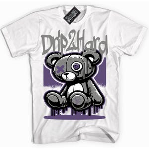 Könnte beinhalten: Weißes T-Shirt mit einem grauen und weißen Cartoon-Teddybär mit einem lila Auge und einem lila X über dem anderen Auge. Der Bär tropft mit lila Farbe. Der Text "Drip2Hard" befindet sich über dem Bären in grauen Buchstaben.