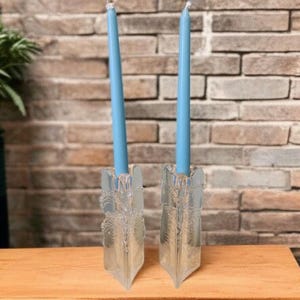 May include: Two clear glass candle holders with blue candles. The candle holders have a decorative, etched design. The candles are a light blue color. The background is a brick wall and a wooden surface.