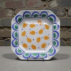 May include: A white, octagonal ceramic serving platter with a hand-painted design. The platter features a repeating pattern of orange fish and blue dots on the interior. The rim is decorated with blue, green, and white scalloped accents. The platter is displayed on a metal stand.
