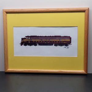 May include: Framed print of a maroon and gold Pennsylvania Railroad diesel locomotive. The artwork is set against a yellow mat and framed in a light brown wooden frame. The locomotive is labeled "Pennsylvania" and "6818".