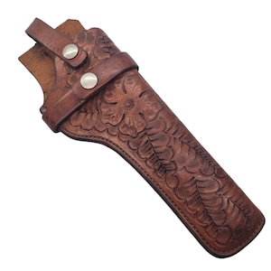 May include: A brown leather holster with floral and leaf tooling. The holster has a strap with two silver-colored snaps. The leather is a medium brown color and appears to be well-worn.