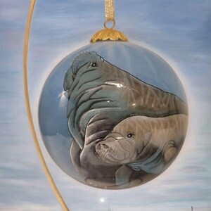 May include: A glass Christmas ornament with a hand-painted image of two manatees, one larger than the other, swimming in a blue and white water background.