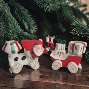 May include: A ceramic train set featuring a white locomotive with a red roof and a Santa Claus figure, followed by a red carriage carrying a stack of white gift boxes with green holly accents. Both train cars have white wheels with gold spokes.