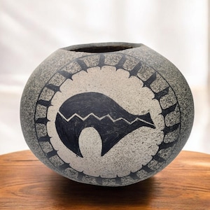 May include: A gray, round ceramic vase with a black and white tribal design. The vase features a central black graphic with a zigzag pattern and a circular border with pointed shapes. The vase sits on a wooden surface.