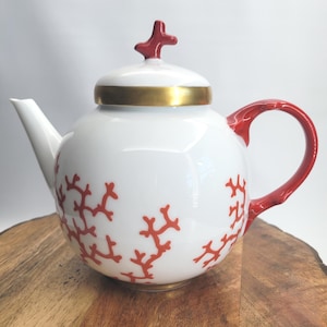 May include: White ceramic teapot with a red coral design. The teapot features a gold band around the lid and a red handle. The lid is topped with a red coral-shaped knob. The teapot is sitting on a wooden surface.