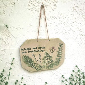 May include: A rectangular ceramic wall plaque with clipped corners, featuring a beige surface with green botanical illustrations of various herbs and the printed text "Friends and Herbs are Everlasting." It hangs from a thin brown twine cord.