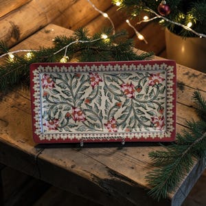 May include: A rectangular ceramic serving tray with a red border and floral design. The tray features a cream-colored background with pink flowers and green leaves. It is displayed on a rustic wooden surface, with Christmas decorations in the background.