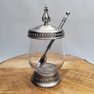 May include: A clear glass sugar bowl with a silver-colored lid, base, and spoon. The lid is topped with a decorative finial. The spoon rests inside the bowl, and the base has a decorative beaded edge. The bowl is on a wooden surface.