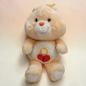 May include: A plush, light-yellow Care Bear with a heart-shaped lock design on its belly. The bear has a white muzzle, a small orange nose, and brown eyes. A small, yellow tuft of hair sits atop its head. The bear is a vintage toy.