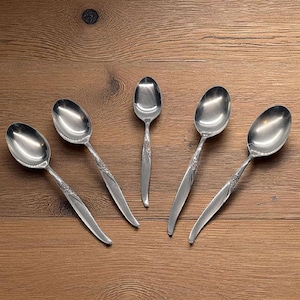 May include: Five silver spoons with a floral design on the handles are arranged on a wooden surface. The spoons have a classic, elegant design, with a polished finish. The spoons are likely for serving or eating.