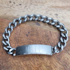 May include: A silver-colored curb chain bracelet with a rectangular nameplate. The name "MATT" is engraved on the nameplate. The bracelet is displayed on a wooden surface.
