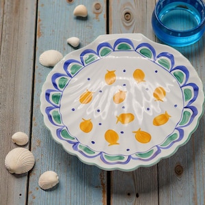 May include: White ceramic seashell-shaped serving dish with a scalloped edge, decorated with blue, green, and yellow accents. The dish features orange fish designs and blue dots. Seashells and a blue glass are nearby.