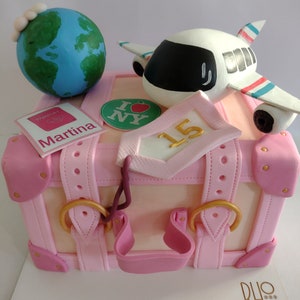 Puede incluir: Un pastel rosa con forma de maleta decorado con elementos temáticos de viaje. Presenta un globo terráqueo, un avión de juguete y un pasaporte. El pastel está adornado con correas rosas, detalles dorados y una etiqueta con el número 15.