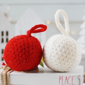May include: Two knitted Christmas ornaments, one red and one white, with a white box that says "PEACE." in red lettering.