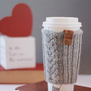 Puede incluir: Un calentador de taza de caf&eacute; de punto gris con una etiqueta de cuero marr&oacute;n que dice "Coffee Queen". El calentador est&aacute; en una taza para llevar blanca con una tapa blanca. La taza est&aacute; sobre un libro marr&oacute;n.