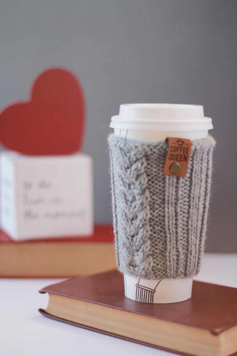 Puede incluir: Un calentador de taza de caf&eacute; de punto gris con una etiqueta de cuero marr&oacute;n que dice "Coffee Queen". El calentador est&aacute; en una taza para llevar blanca con una tapa blanca. La taza est&aacute; sobre un libro marr&oacute;n.