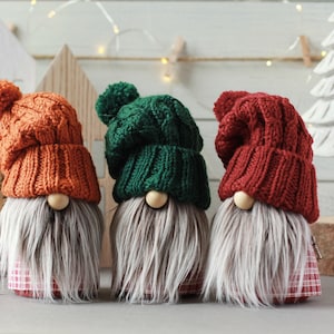 Op de afbeelding: Drie gebreide gnomen met verschillende gekleurde mutsen. De gnome links heeft een oranje muts, de middelste gnome een groene muts en de gnome rechts een rode muts. Alle drie de gnomen hebben witte baarden en dragen geruite shirts.