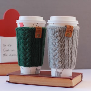 Puede incluir: Dos tazas de caf&eacute; con mangas de punto. La manga verde tiene una etiqueta de cuero marr&oacute;n con el texto "SWHHH COFFEE". La manga gris tiene una etiqueta de cuero marr&oacute;n con el texto "COFFEE QUEEN".