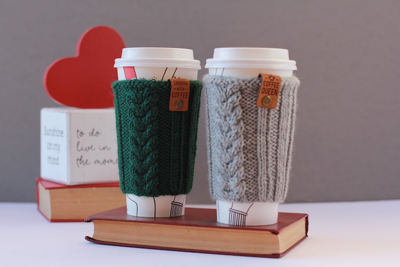 Puede incluir: Dos tazas de caf&eacute; con mangas de punto. La manga verde tiene una etiqueta de cuero marr&oacute;n con el texto "SWHHH COFFEE". La manga gris tiene una etiqueta de cuero marr&oacute;n con el texto "COFFEE QUEEN".