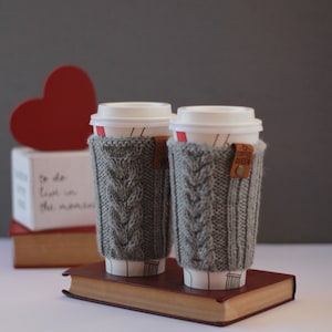 Puede incluir: Dos fundas de caf&eacute; de punto gris con una etiqueta de cuero marr&oacute;n que dice "Coffee Queen" en un libro marr&oacute;n. Las fundas est&aacute;n encima de dos tazas de caf&eacute; blancas.
