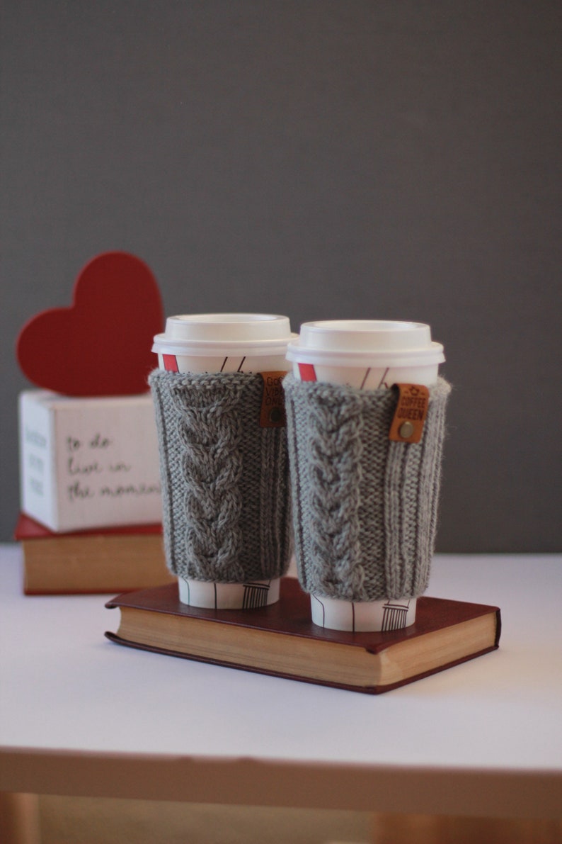 Puede incluir: Dos fundas de caf&eacute; de punto gris con una etiqueta de cuero marr&oacute;n que dice "Coffee Queen" en un libro marr&oacute;n. Las fundas est&aacute;n encima de dos tazas de caf&eacute; blancas.