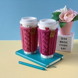 Puede incluir: Dos fundas de taza de caf&eacute; de punto rosa con una etiqueta marr&oacute;n que dice "tea guild" en una libreta azul con un bol&iacute;grafo. La libreta est&aacute; sobre una superficie amarilla. Una maceta rosa con flores blancas est&aacute; en el fondo.