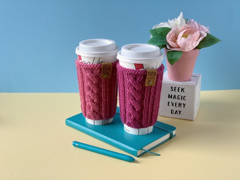 Puede incluir: Dos fundas de taza de caf&eacute; de punto rosa con una etiqueta marr&oacute;n que dice "tea guild" en una libreta azul con un bol&iacute;grafo. La libreta est&aacute; sobre una superficie amarilla. Una maceta rosa con flores blancas est&aacute; en el fondo.