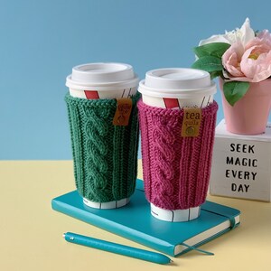 Puede incluir: Dos tazas de caf&eacute; con mangas de punto verde y rosa. Las mangas tienen un patr&oacute;n de punto de cable. Las tazas est&aacute;n sobre un cuaderno azul con un bol&iacute;grafo. El fondo es una pared azul claro y una superficie amarilla. Hay una maceta rosa con una flor blanca sobre un cubo blanco con el texto "SEEK MAGIC EVERY DAY".