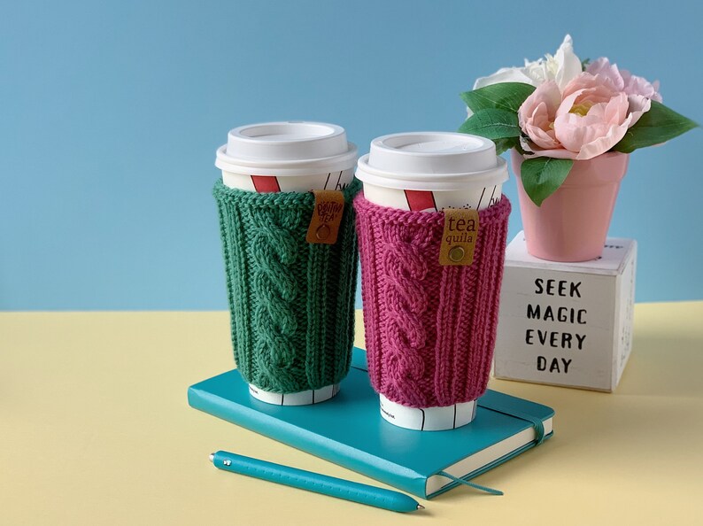 Puede incluir: Dos tazas de caf&eacute; con mangas de punto verde y rosa. Las mangas tienen un patr&oacute;n de punto de cable. Las tazas est&aacute;n sobre un cuaderno azul con un bol&iacute;grafo. El fondo es una pared azul claro y una superficie amarilla. Hay una maceta rosa con una flor blanca sobre un cubo blanco con el texto "SEEK MAGIC EVERY DAY".